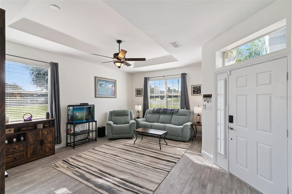 A living room features a tray ceiling, light wood-look flooring, a sofa, and a recliner — 2720 SAVANNAH DRIVE, LEESBURG, FL