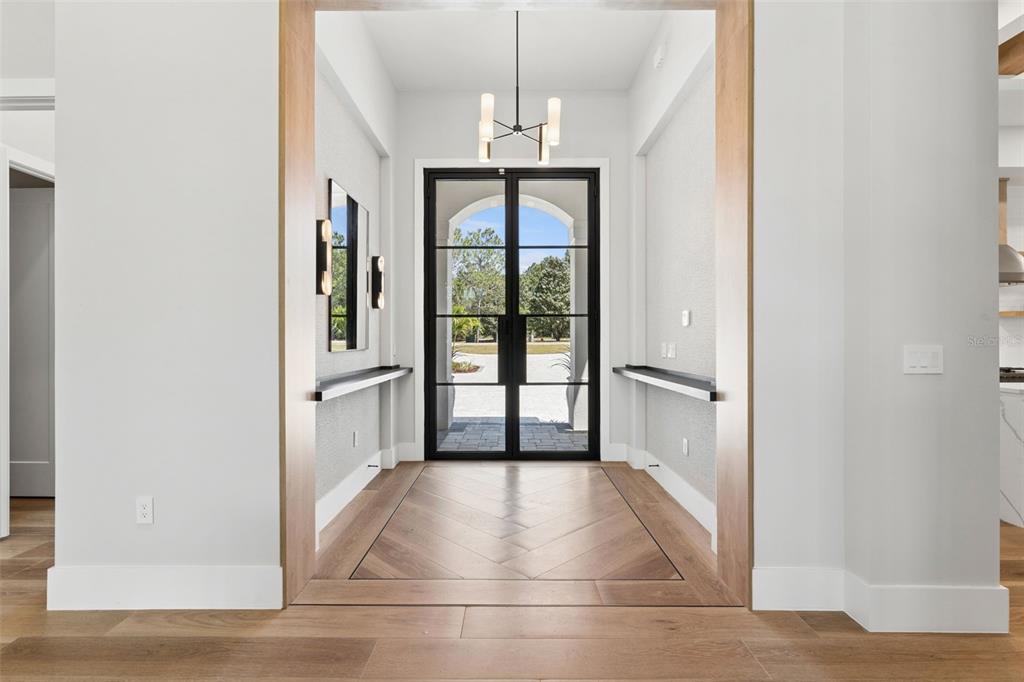 A modern entryway features a black-framed glass double door, herringbone wood flooring, and wall-mounted consoles — 16989 LUCCA LANE, MONTVERDE, FL