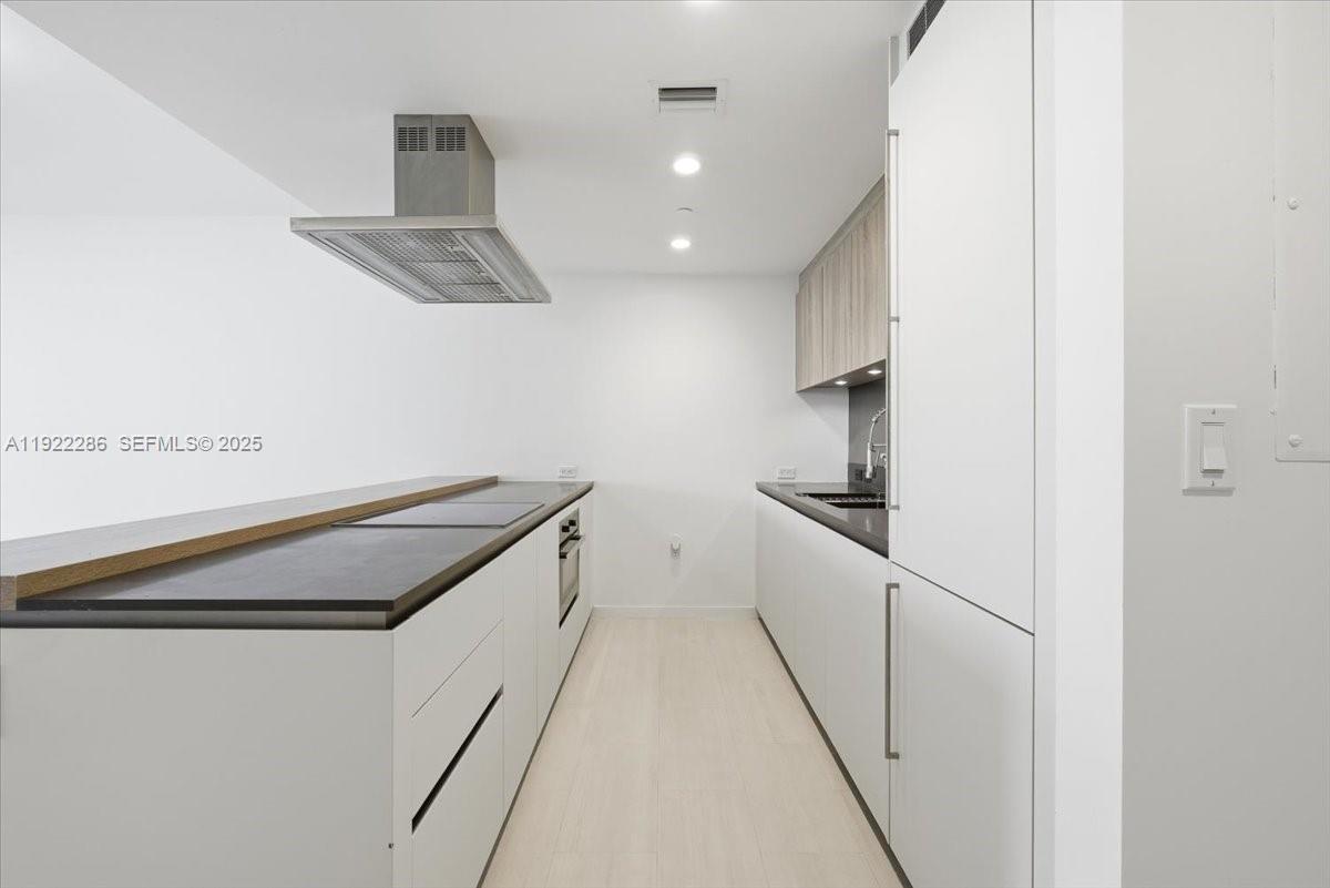 A galley-style kitchen features white cabinetry, dark countertops, and a stainless steel range hood — 1000 Brickell Plz # 3211, Miami FL 33131, MIAMI, FL