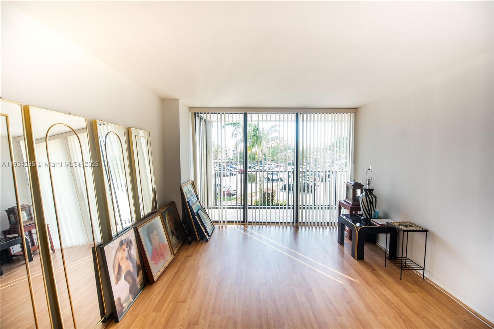 An empty room features wood-look flooring, large gold-framed mirrors, framed artwork, and vertical window blinds — 200 Leslie Dr # 214, Hallandale Beach FL 33009, HALLANDALE BEACH, FL