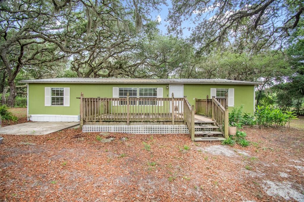 A green single-wide mobile home features a wooden deck and stairs under large oak trees — 156 KINGFISH ROAD, POMONA PARK, FL
