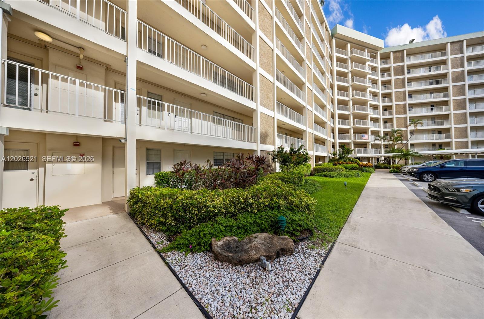 A multi-story apartment building features white balconies, a landscaped garden, and a paved parking area — POMPANO BEACH, FL