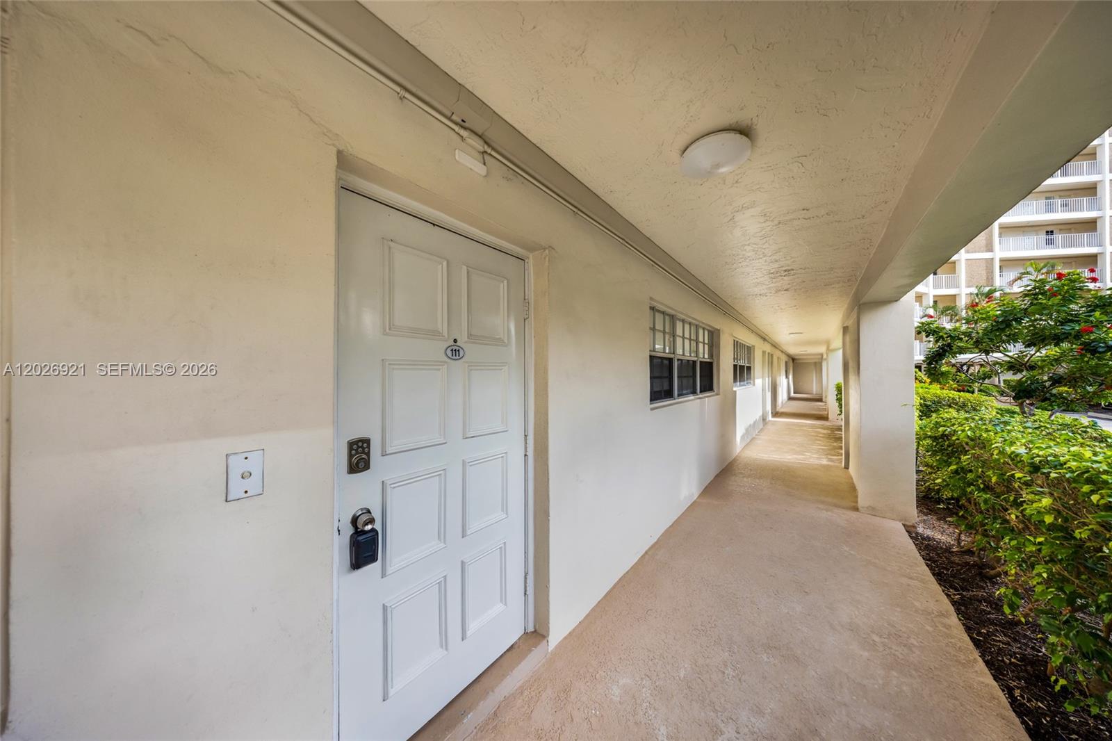 A white door marked 111 opens onto an outdoor walkway lined with green bushes — POMPANO BEACH, FL