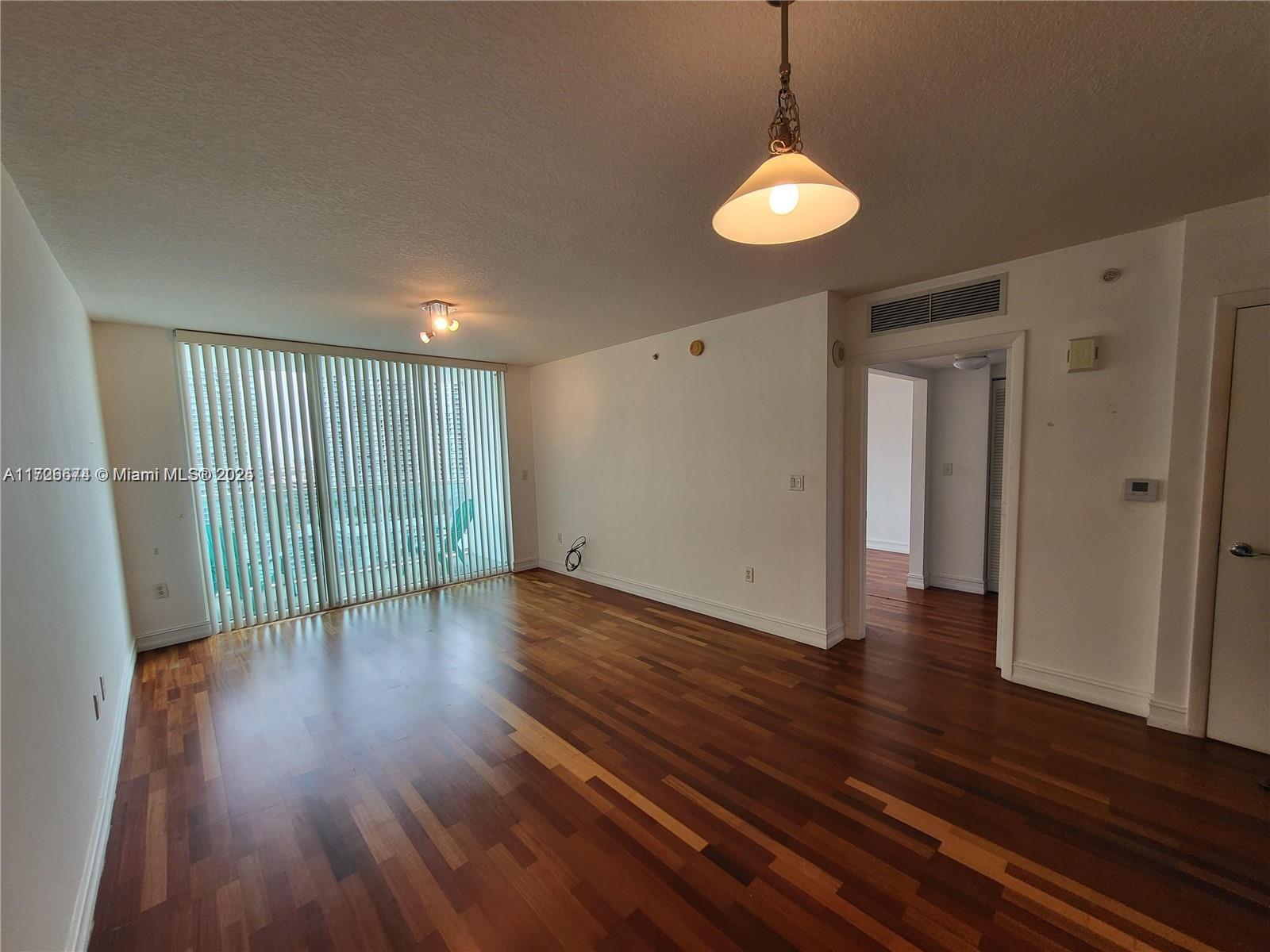 An empty living room features dark hardwood floors, a pendant light, and floor-to-ceiling vertical blinds — 31 SE 5th St # 2409, Miami FL 33131, MIAMI, FL