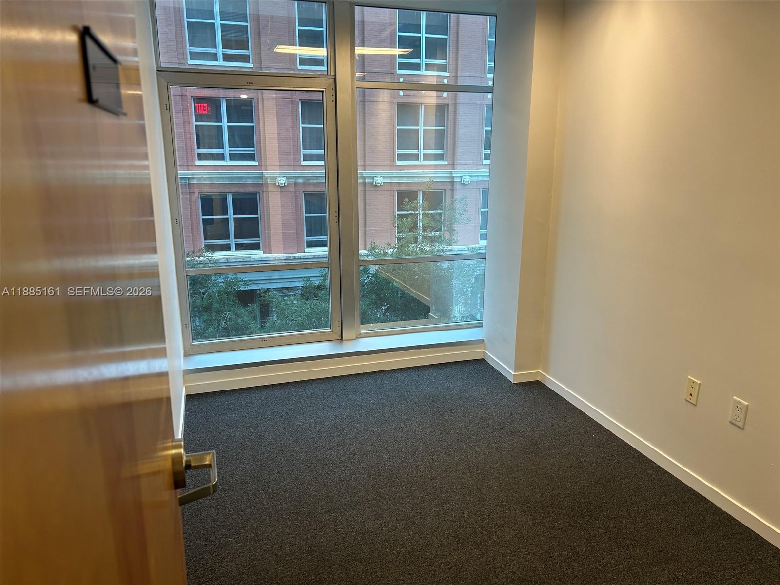 A small office room features dark gray carpeting, white walls, and large windows overlooking buildings — 14 NE 1st Ave # 500D, Miami FL 33132, MIAMI, FL