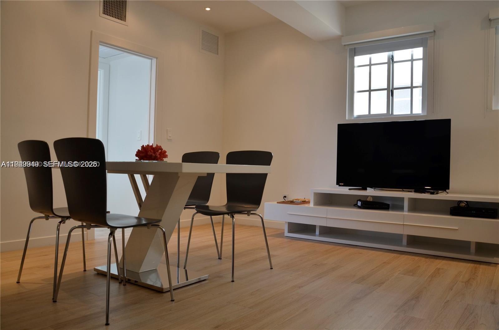 A dining area with a white table and four chairs sits before a television console — 700 16th St # 201, Miami Beach FL 33139, MIAMI BEACH, FL