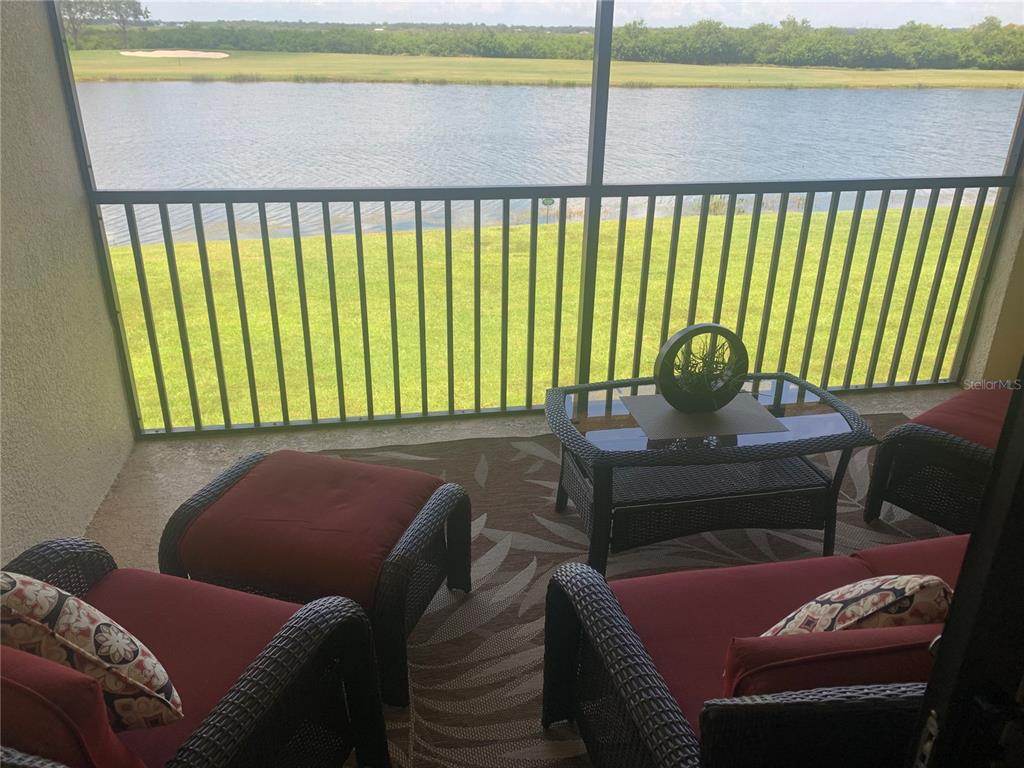 A screened-in patio features wicker furniture with red cushions overlooking a lake and golf course — 7225 RIVER HAMMOCK DRIVE UNIT 202, BRADENTON, FL