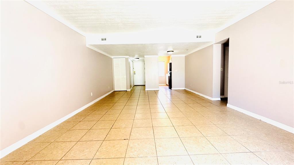 An empty room with light-colored walls and tan tile flooring leads toward a kitchen area — 5508 POKEWEED COURT UNIT 146, TAMPA, FL