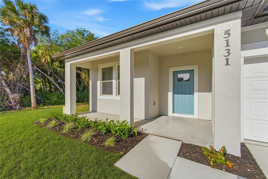 A light-colored house exterior features a blue front door, a covered porch, and landscaped garden — 5131 S SAN MATEO DRIVE, NORTH PORT, FL