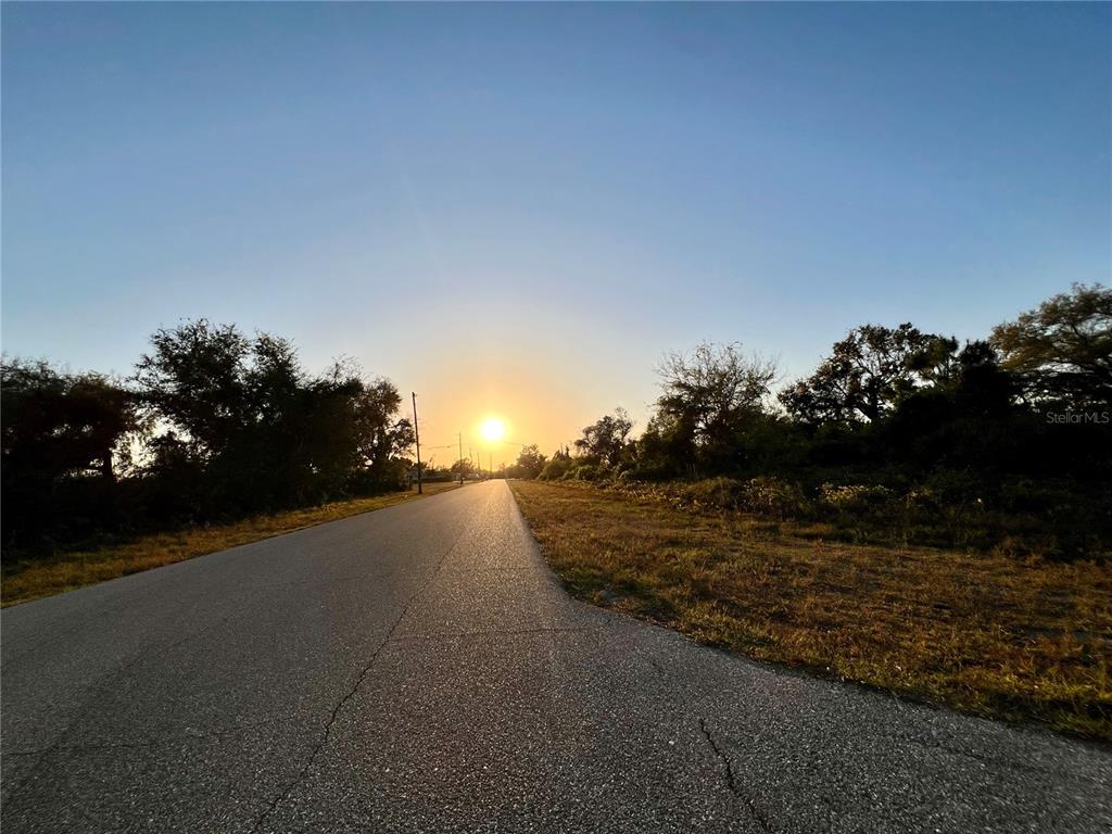 A paved road stretches toward a bright sunset between trees and grassy fields — 12332 ALBRECHT TERRACE, PORT CHARLOTTE, FL