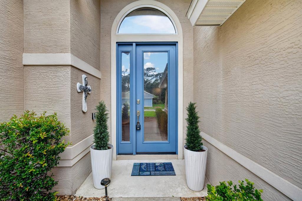 A blue front door with a glass sidelight and arched transom sits between two potted plants — 1234 CROWN POINTE LANE, ORMOND BEACH, FL