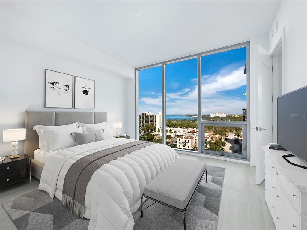 A modern bedroom features a bed, a bench, and floor-to-ceiling windows overlooking a coastal city — 301 QUAY COMMONS UNIT 1003, SARASOTA, FL