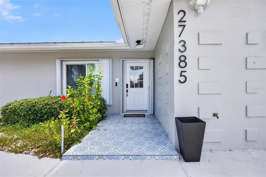 The exterior of a house features a white front door, patterned tile entryway, and address numbers — 27385 VOYAGEUR DRIVE, PUNTA GORDA, FL
