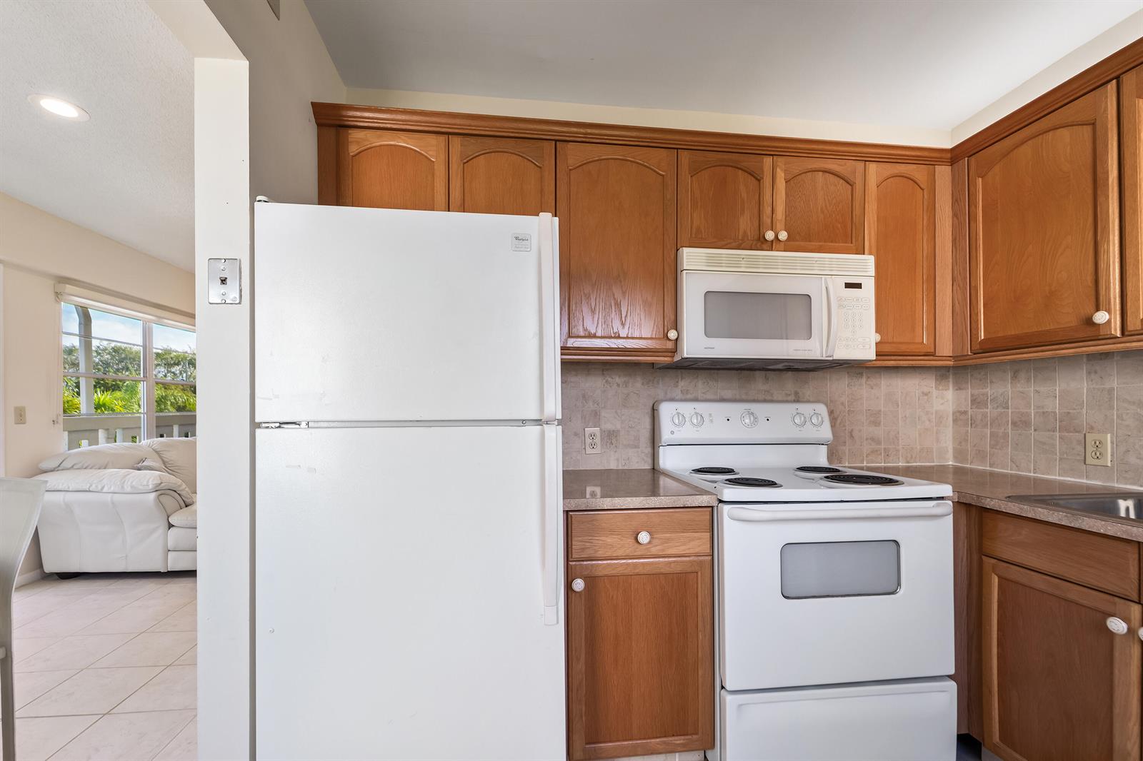 A kitchen features wood cabinets, white appliances, and a tiled backsplash next to a living area — 200 Dorset E, Boca Raton FL 33434, BOCA RATON, FL