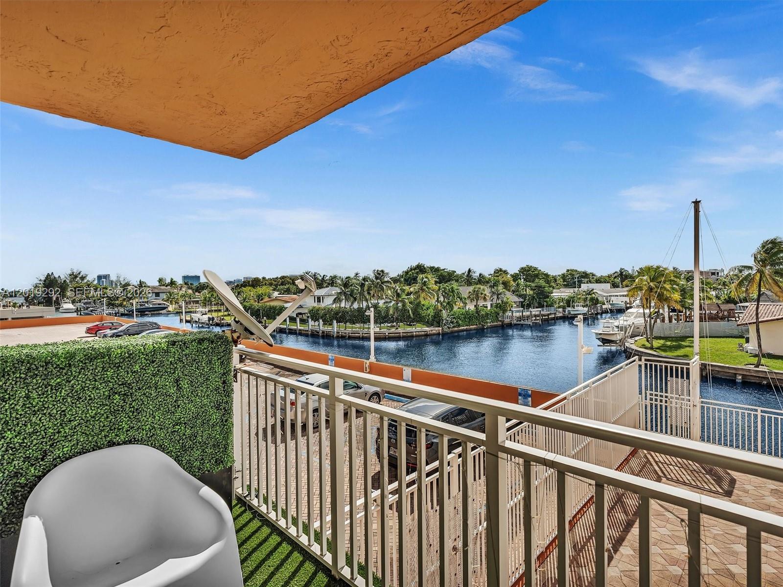 A balcony with a white chair overlooks a canal, parked cars, and palm trees — 2450 NE 135th St # 201, North Miami FL 33181, NORTH MIAMI, FL
