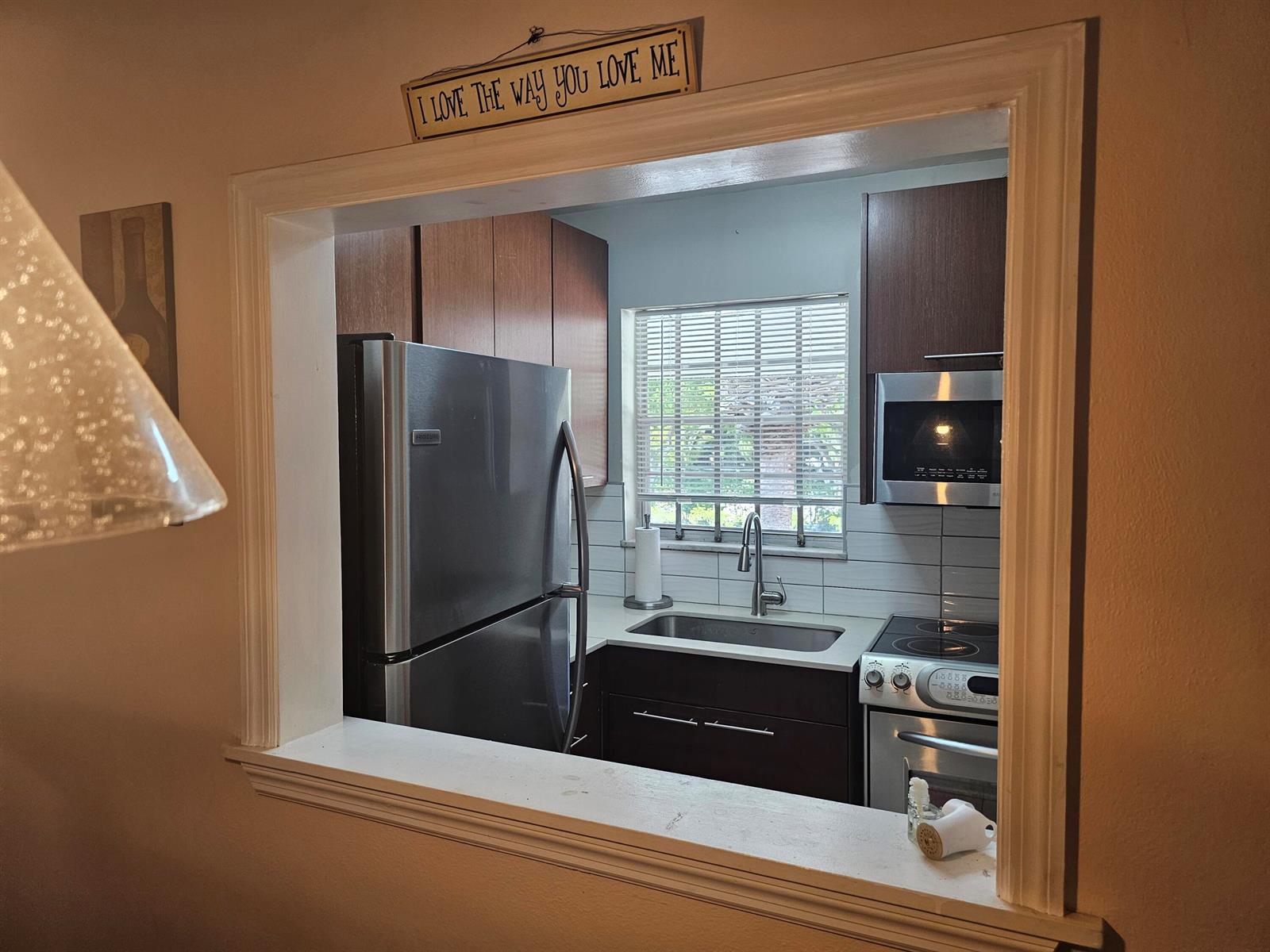 A kitchen viewed through a pass-through window features stainless steel appliances and dark wood cabinetry — 2475 W Golf Boulevard # 243, Pompano Beach FL 33064, POMPANO BEACH, FL
