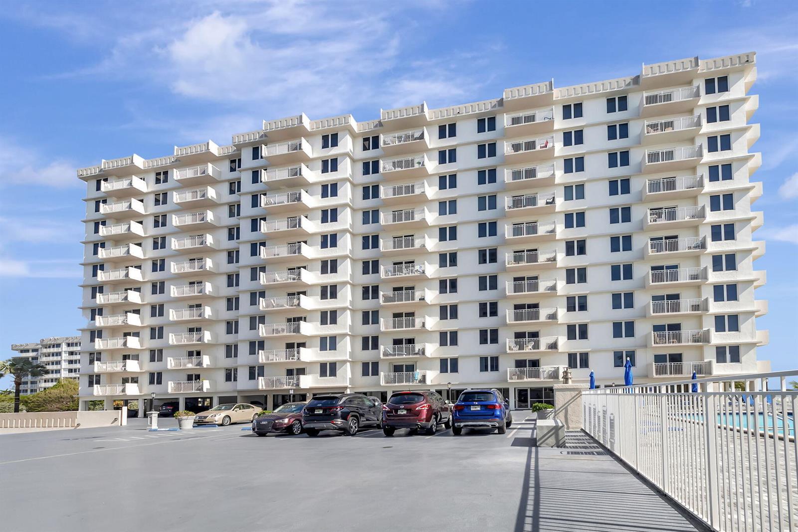 A multi-story white residential building with balconies stands above a paved parking lot — 3215 S Ocean Boulevard # 509, Highland Beach FL 33487, HIGHLAND BEACH, FL
