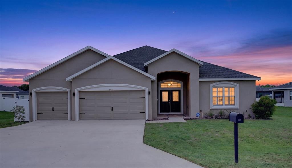 A single-story suburban house with a three-car garage and a paved driveway at sunset — 9074 SE 48TH COURT ROAD, OCALA, FL