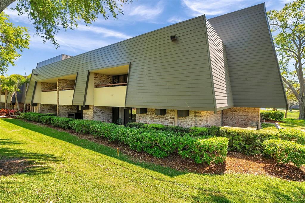 The exterior of a multi-story building features olive-green siding, brick accents, and a landscaped lawn — 36750 US 19 N UNIT 2763, PALM HARBOR, FL