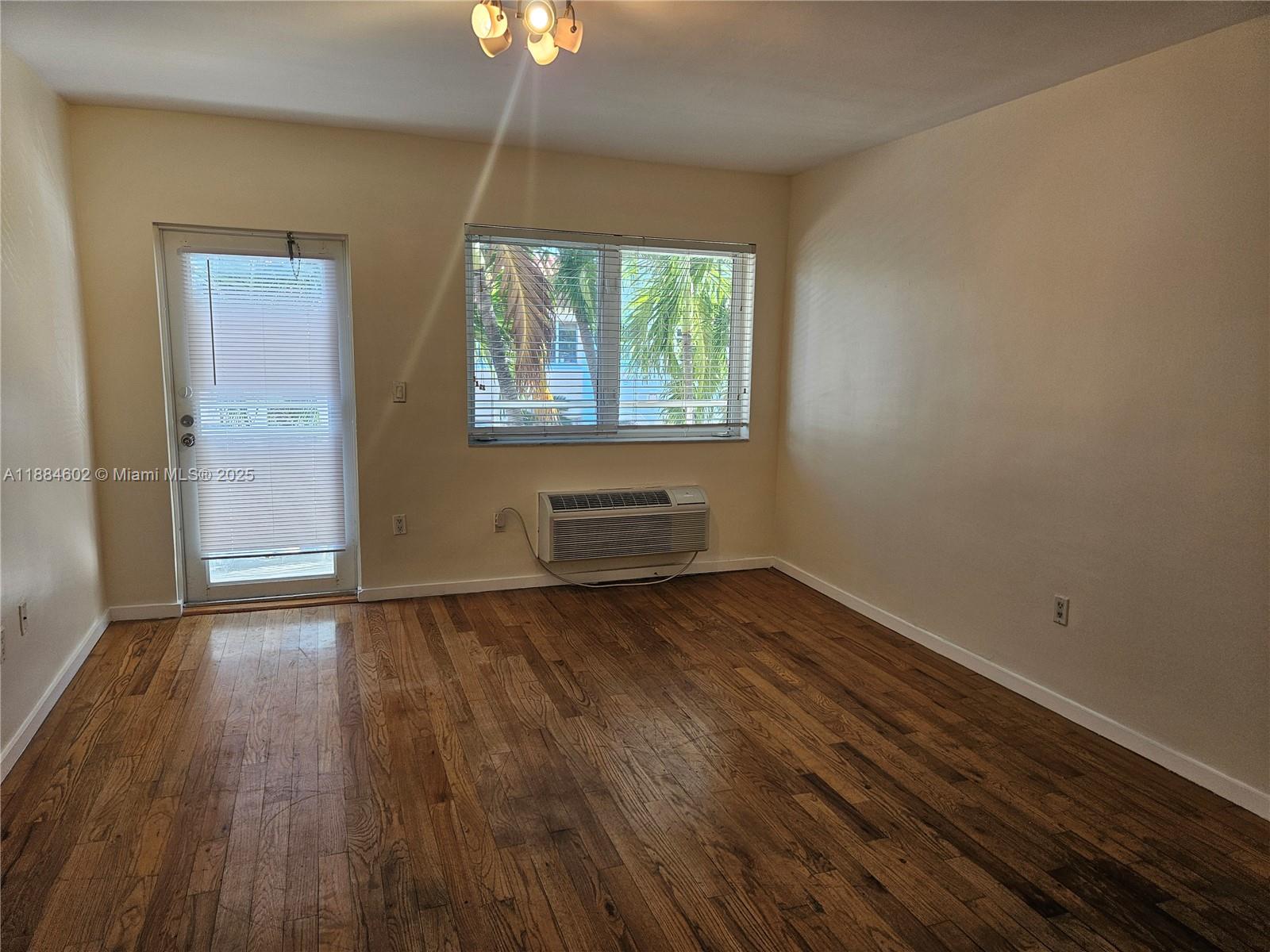 An empty room features hardwood flooring, a window with blinds, a door, and an air conditioner — 745 13th St # 16, Miami Beach FL 33139, MIAMI BEACH, FL