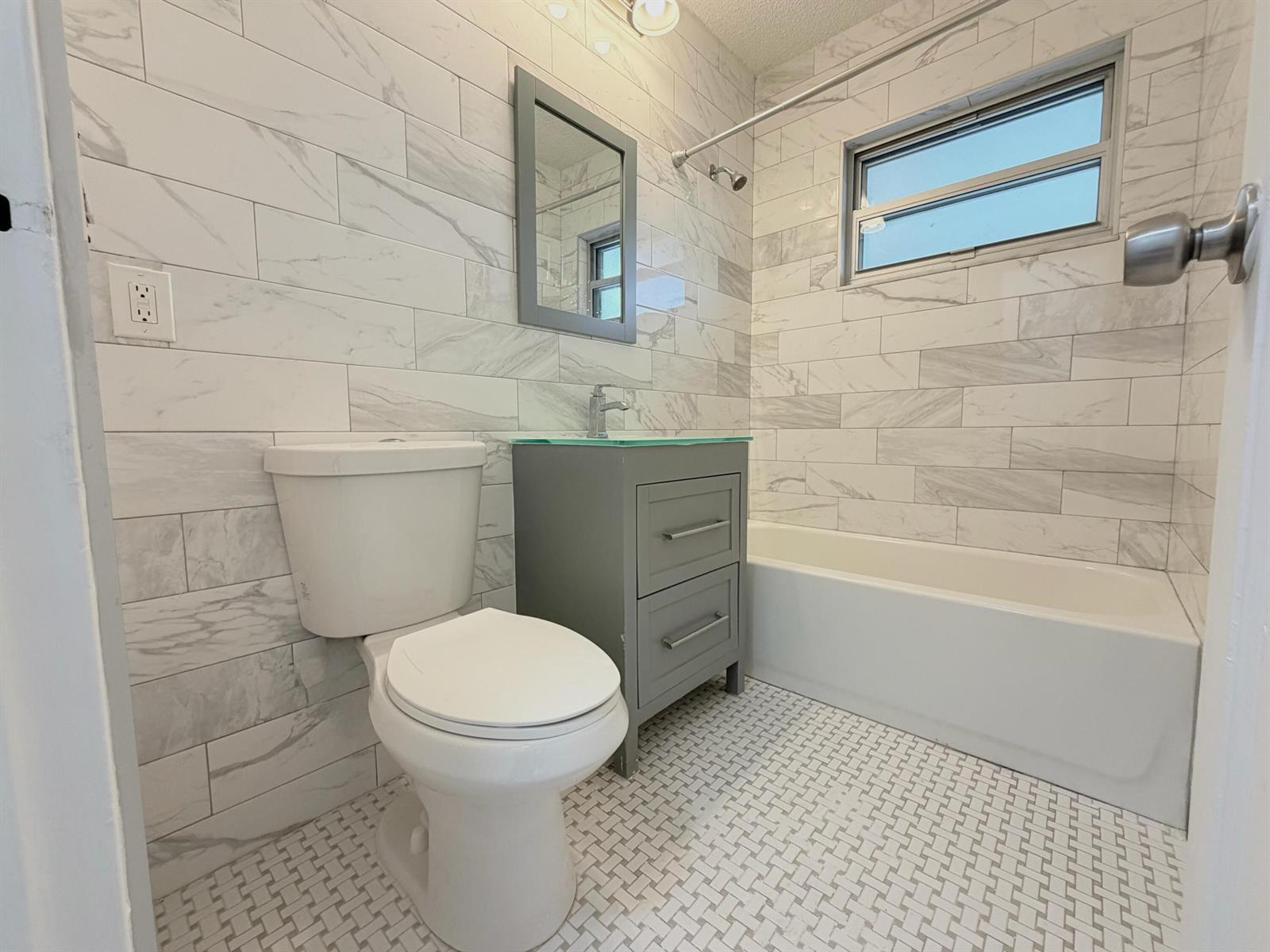 A bathroom features a gray vanity, a white toilet, and a tub with marble-look tiles — 831 El Prado, West Palm Beach FL 33405, WEST PALM BEACH, FL