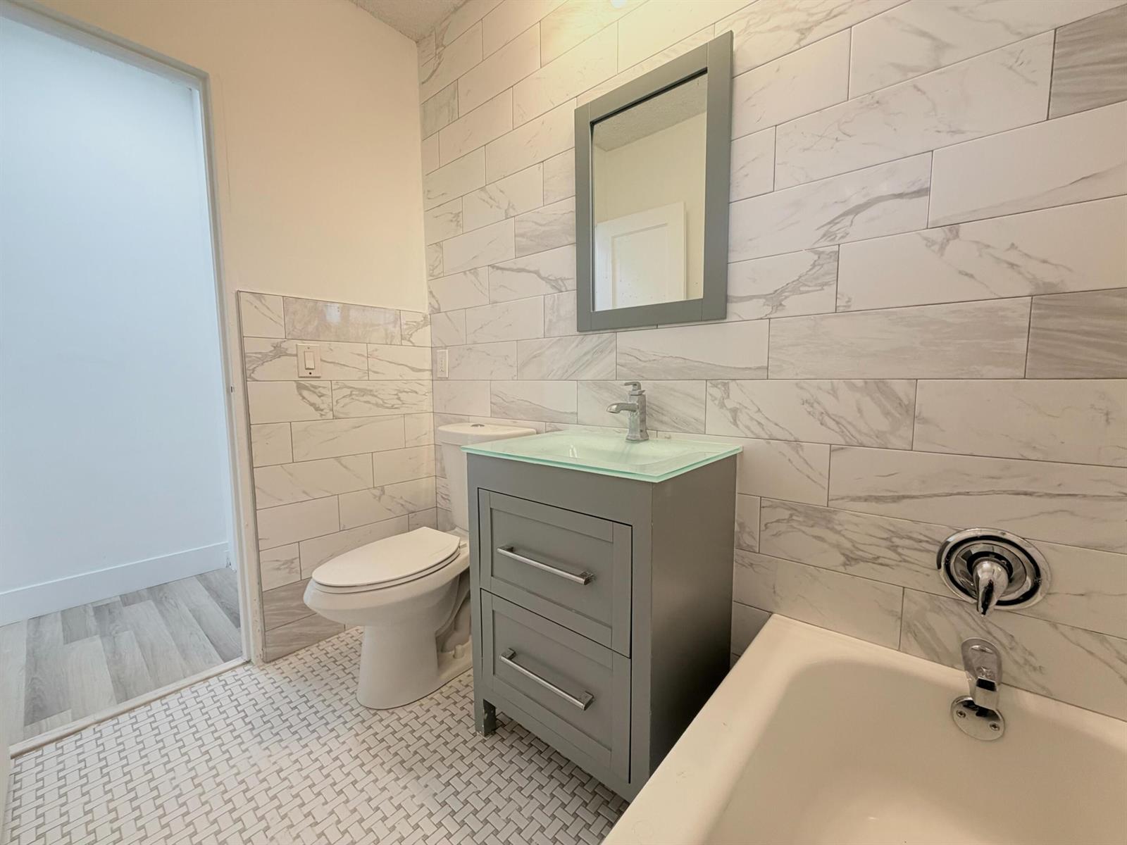 A bathroom features a gray vanity, a glass sink, a toilet, and marble-look wall tiles — 831 El Prado, West Palm Beach FL 33405, WEST PALM BEACH, FL