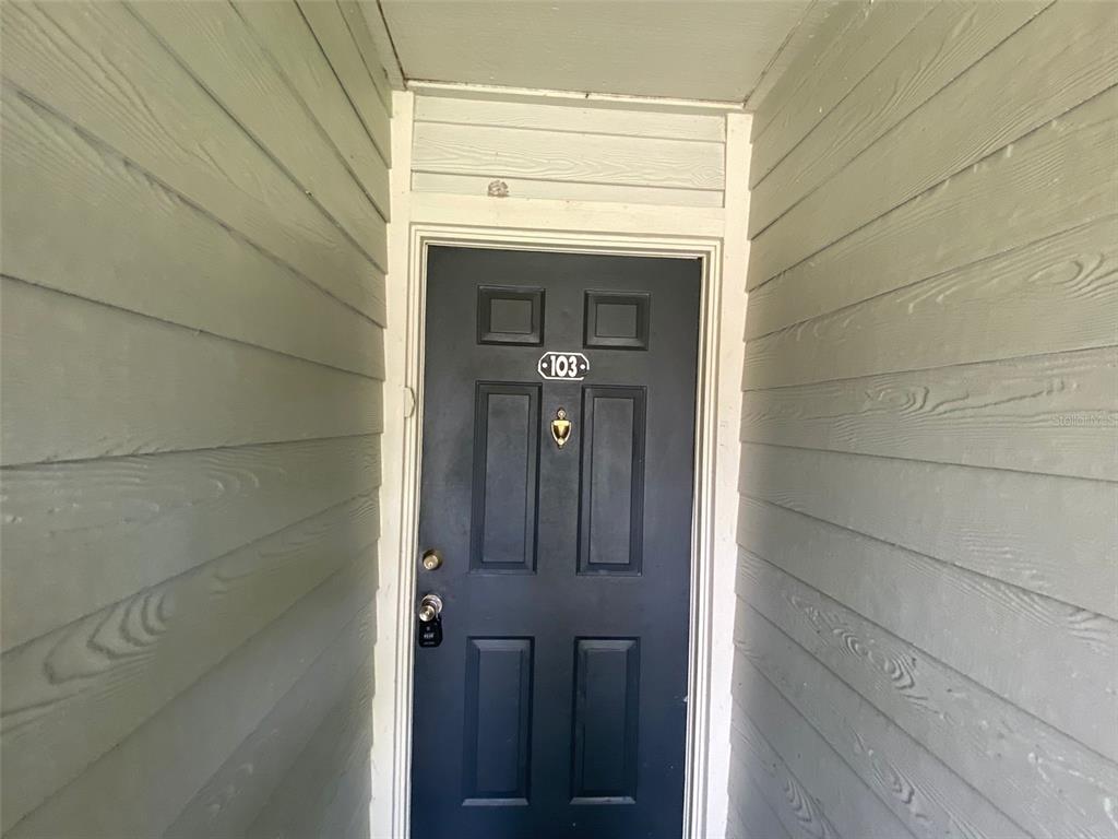 A dark blue front door with the number 103 is framed by light gray siding — 6100 WESTGATE DRIVE UNIT 103, ORLANDO, FL