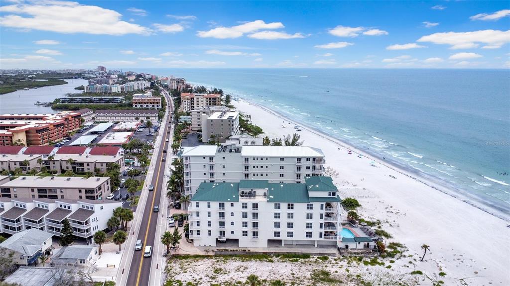 An aerial view shows a coastal road running alongside multi-story apartment buildings and a beach — 19734 GULF BOULEVARD UNIT 402, INDIAN SHORES, FL
