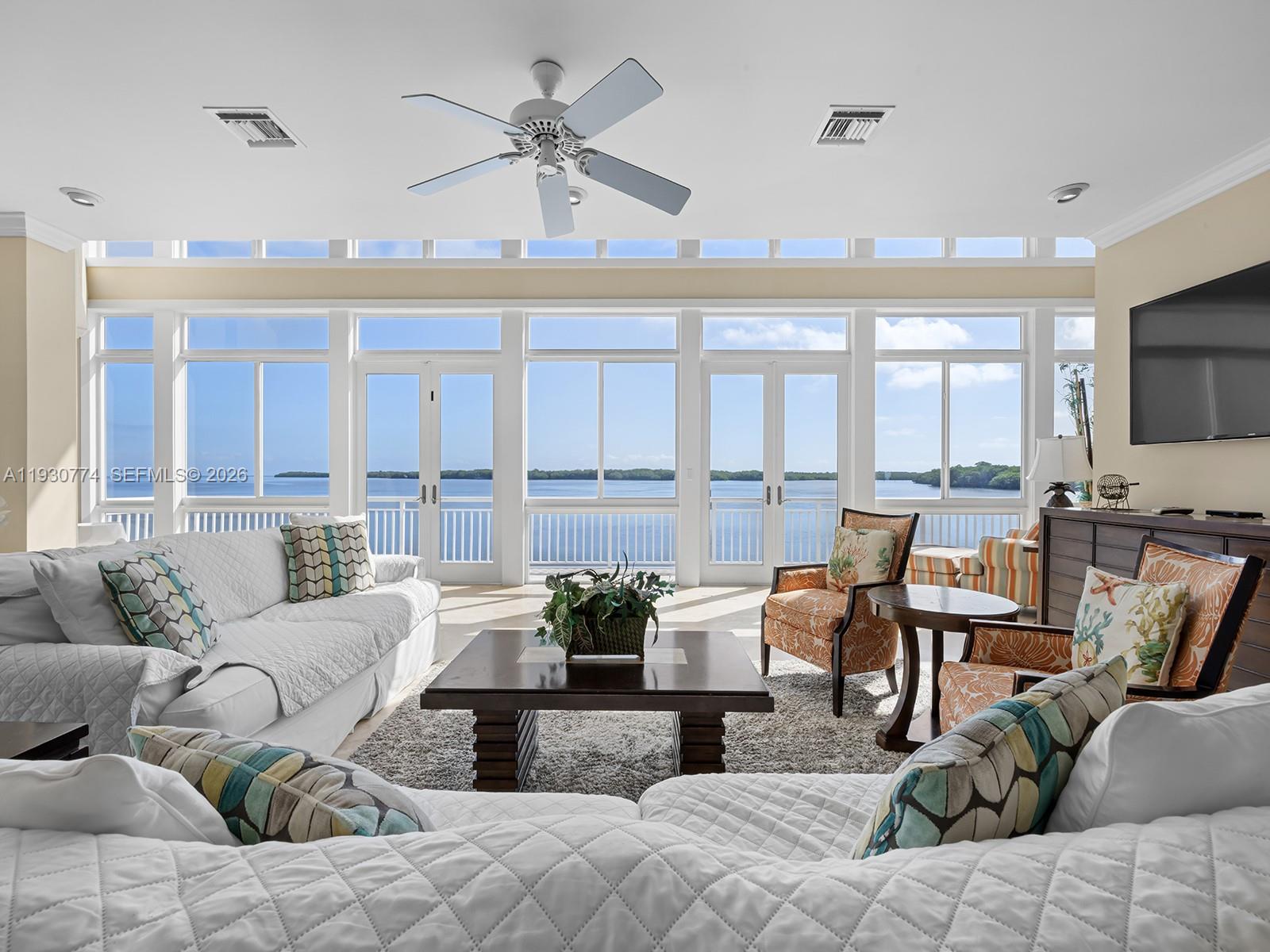 A living room with white sofas, patterned pillows, and large windows overlooking a body of water — 57 Garden Cove Dr, Key Largo FL 33037, KEY LARGO, FL