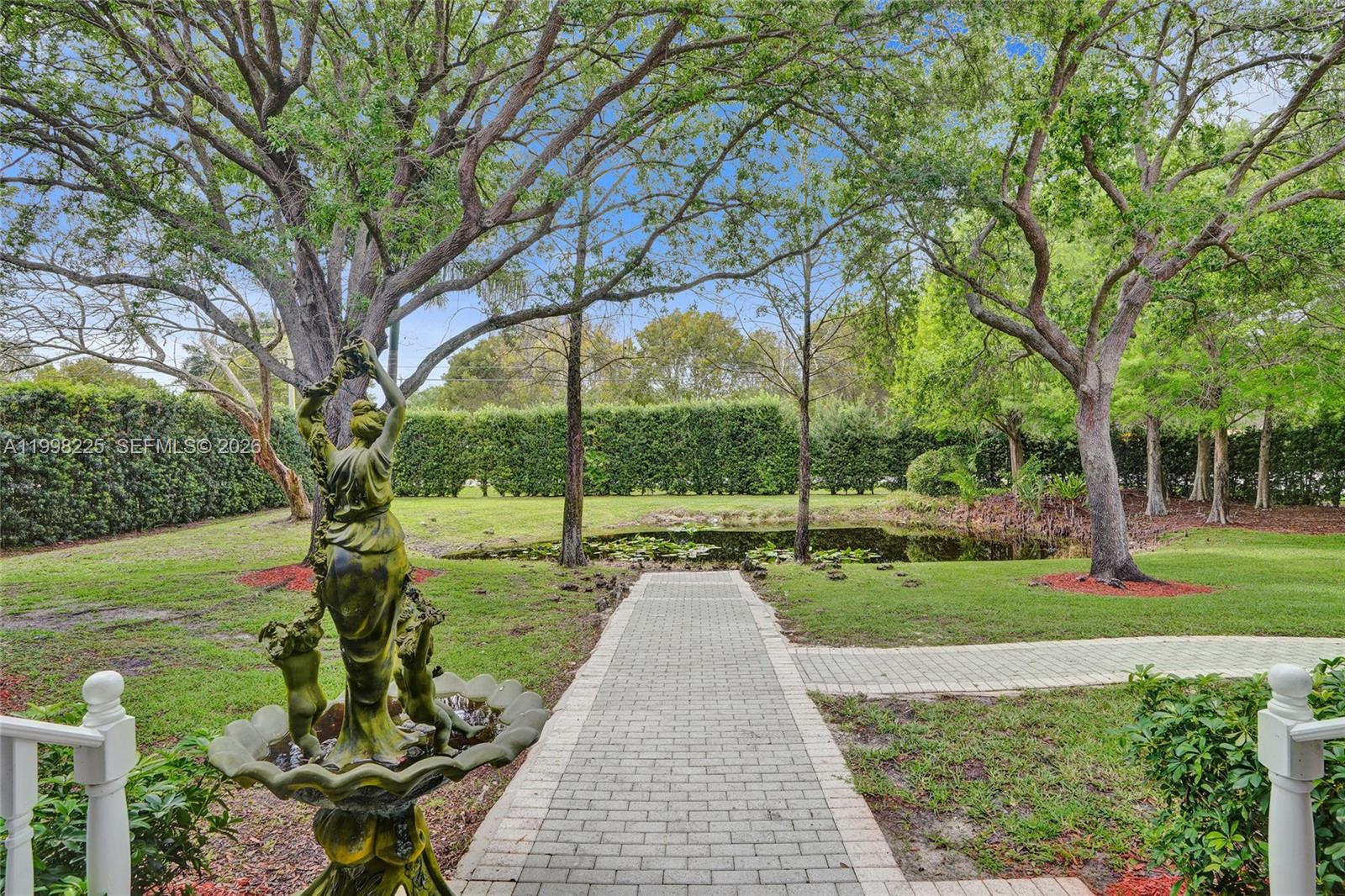 A stone walkway leads toward a small pond in a grassy backyard with mature trees — 12350 NW 6th St, Plantation FL 33325, PLANTATION, FL