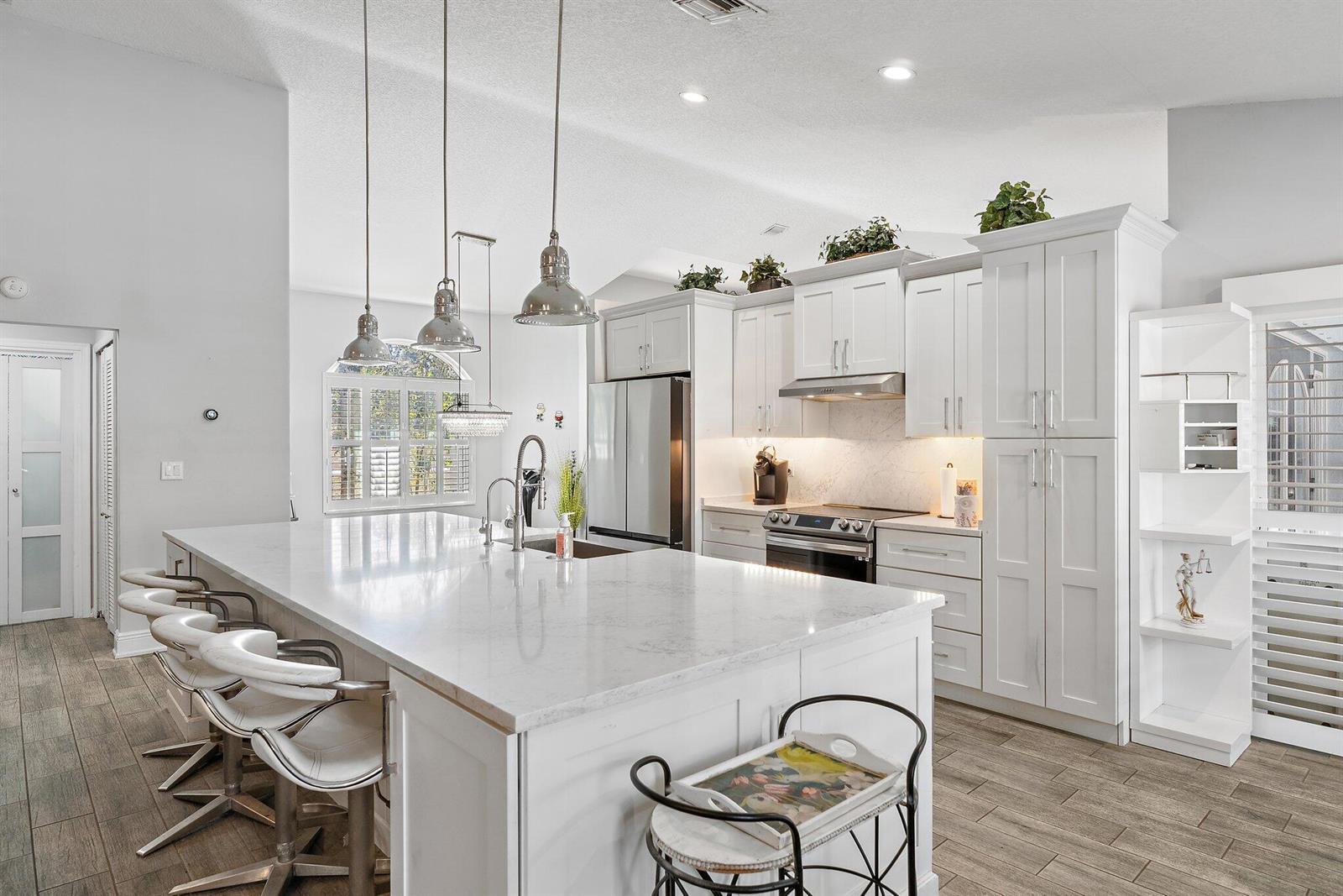 A modern white kitchen features a large quartz island, stainless steel appliances, and pendant lighting — 13033 Randolph Siding Road N, Jupiter FL 33478, JUPITER, FL