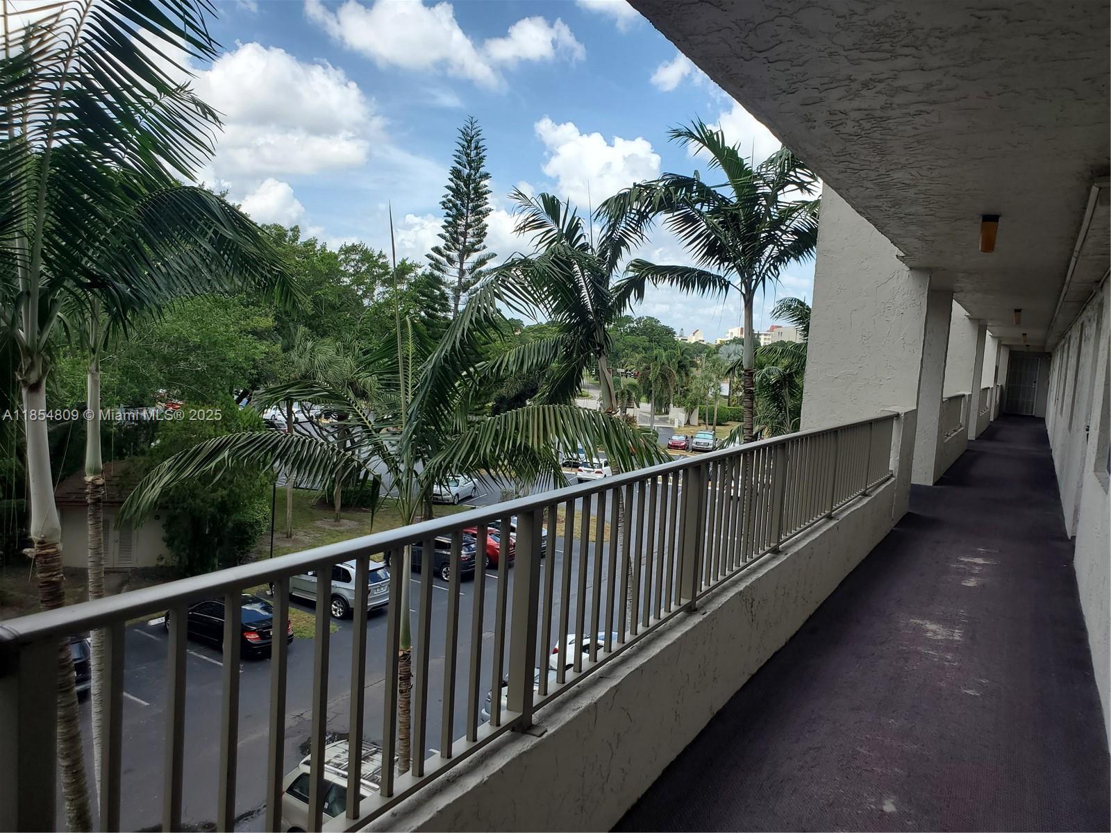 An outdoor apartment walkway overlooks a parking lot with palm trees under a cloudy sky — 3940 Inverrary Blvd # A, Lauderhill FL 33319, LAUDERHILL, FL