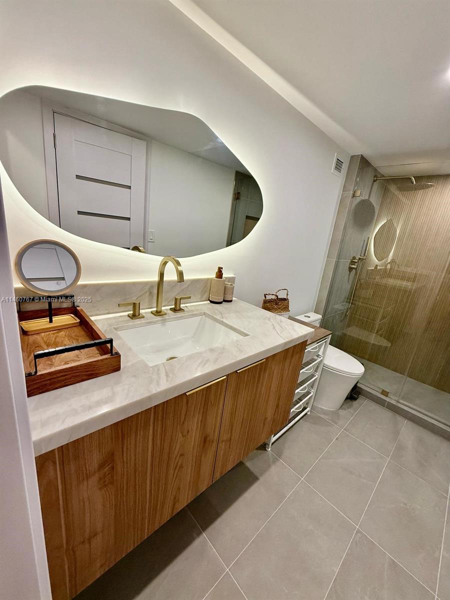 A modern bathroom features a wood vanity, marble countertop, gold faucet, and an illuminated mirror — 2301 Collins Ave # 1121, Miami Beach FL 33139, MIAMI BEACH, FL