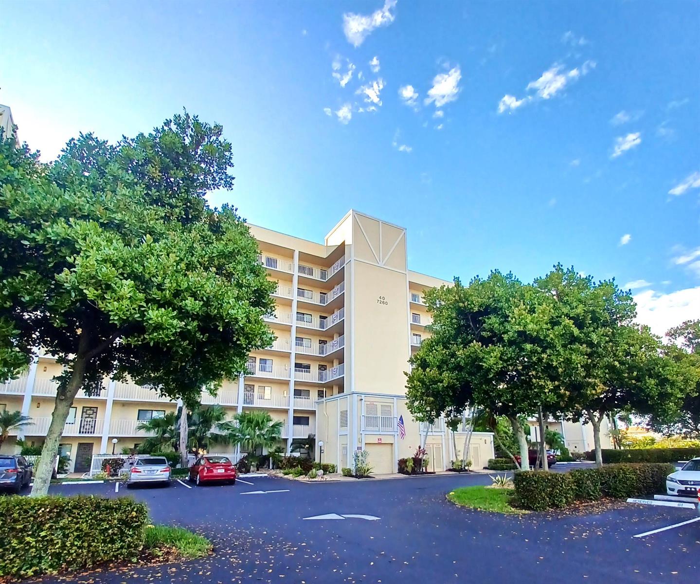 A multi-story apartment building with a parking lot and trees under a blue sky — 7260 Kinghurst Drive # 406, Delray Beach FL 33446, DELRAY BEACH, FL
