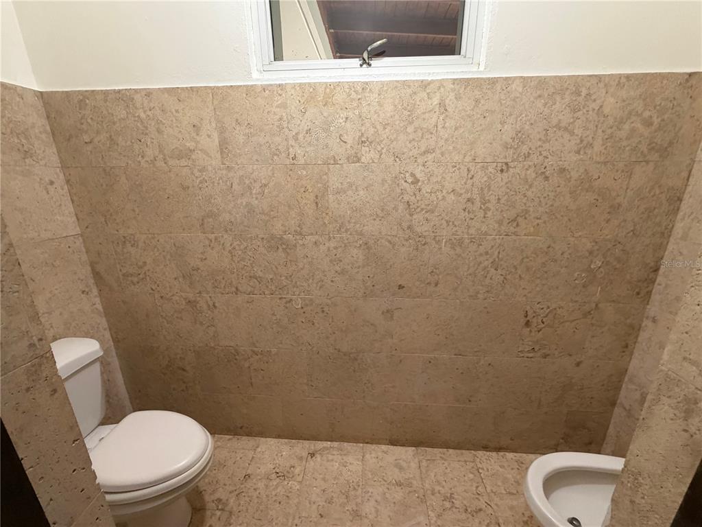 A bathroom features a toilet, a bidet, and walls and floors covered in tan stone tiles — Lot 119 VILLA SONSIRE, MAYAGUEZ, PR