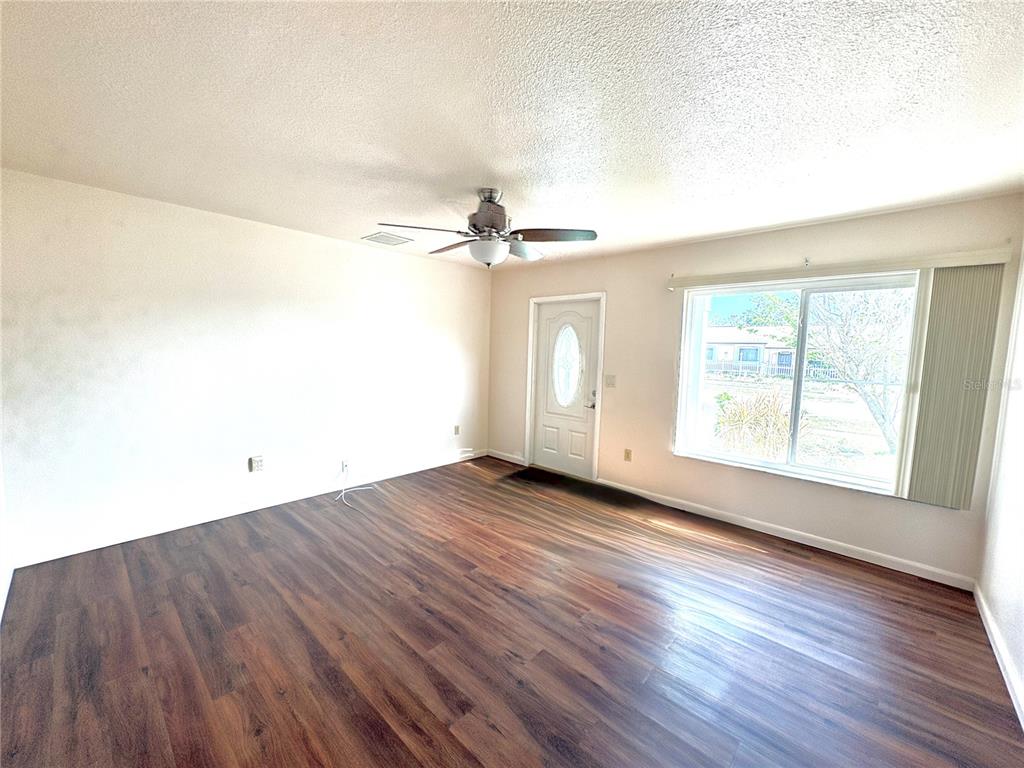 An empty room features dark wood-look flooring, white walls, a ceiling fan, and a front door — 6752 DENNISON AVENUE, NORTH PORT, FL