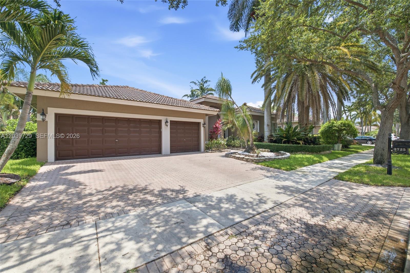 A tan house with a three-car garage and a paved driveway sits under palm trees — 3725 Oak Ridge Ln, Weston FL 33331, WESTON, FL