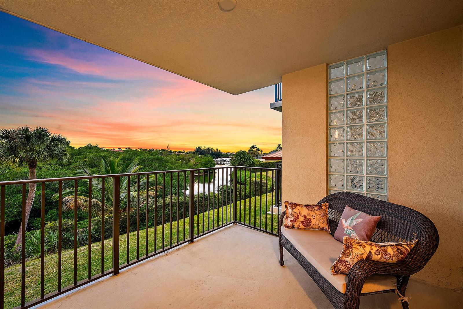 A balcony with a wicker bench overlooks a canal and lush greenery at sunset — 1701 Marina Isle Way # 202, Jupiter FL 33477, JUPITER, FL