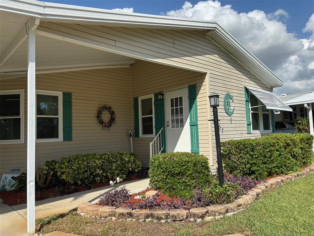 A tan mobile home features a covered porch, green shutters, and a landscaped front garden — 6107 GRAND OAKS DRIVE SE, WINTER HAVEN, FL