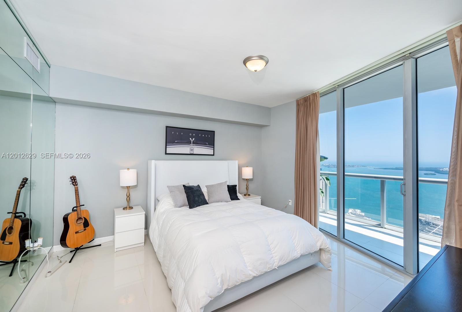 A bedroom features a white bed, two nightstands, a guitar, and floor-to-ceiling windows overlooking water — 465 E Brickell Ave # 5305, Miami FL 33131, MIAMI, FL