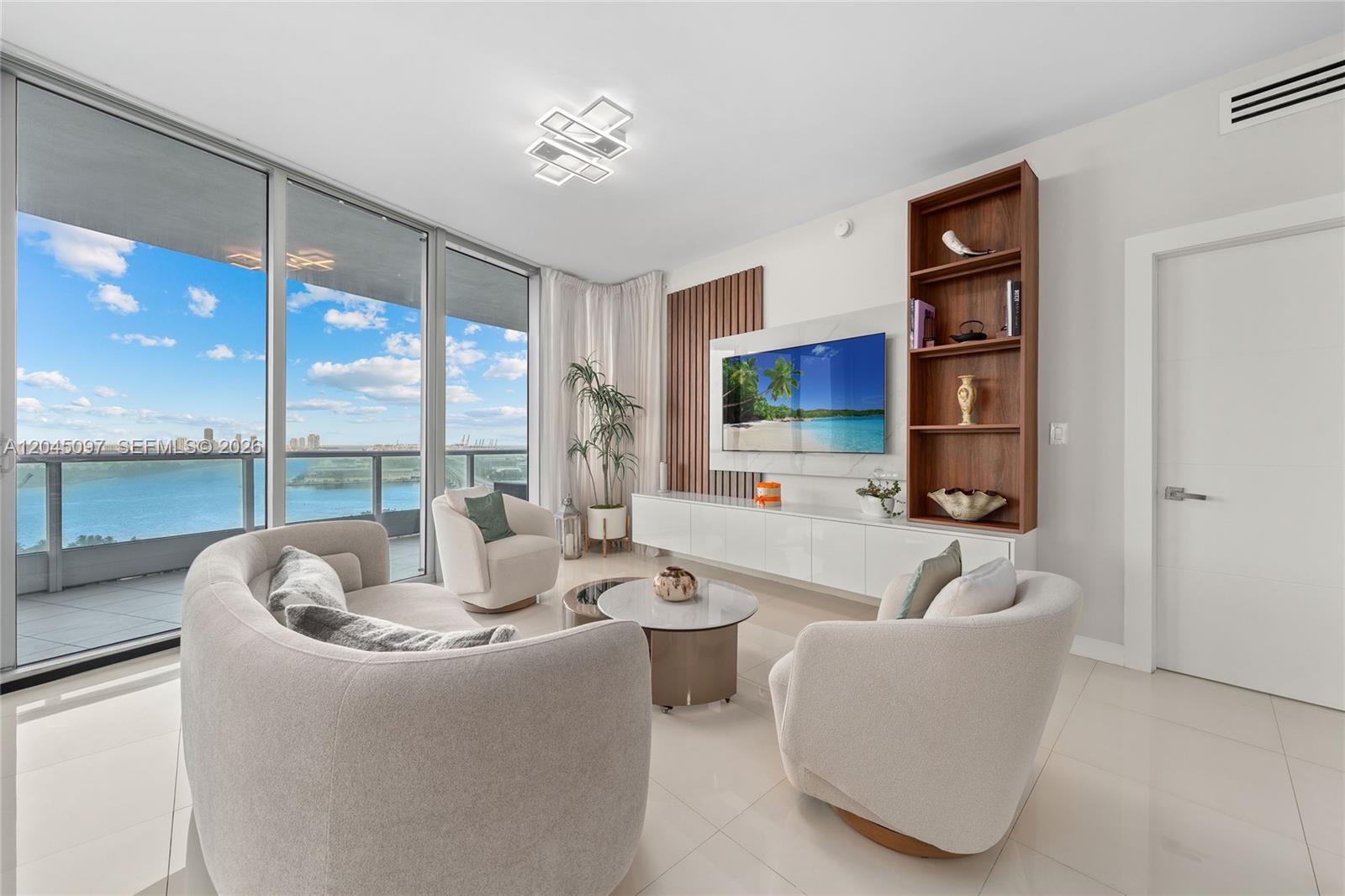 A modern living room features curved seating, a wall-mounted television, and floor-to-ceiling windows overlooking water — 900 Biscayne Blvd # 2506, Miami FL 33132, MIAMI, FL