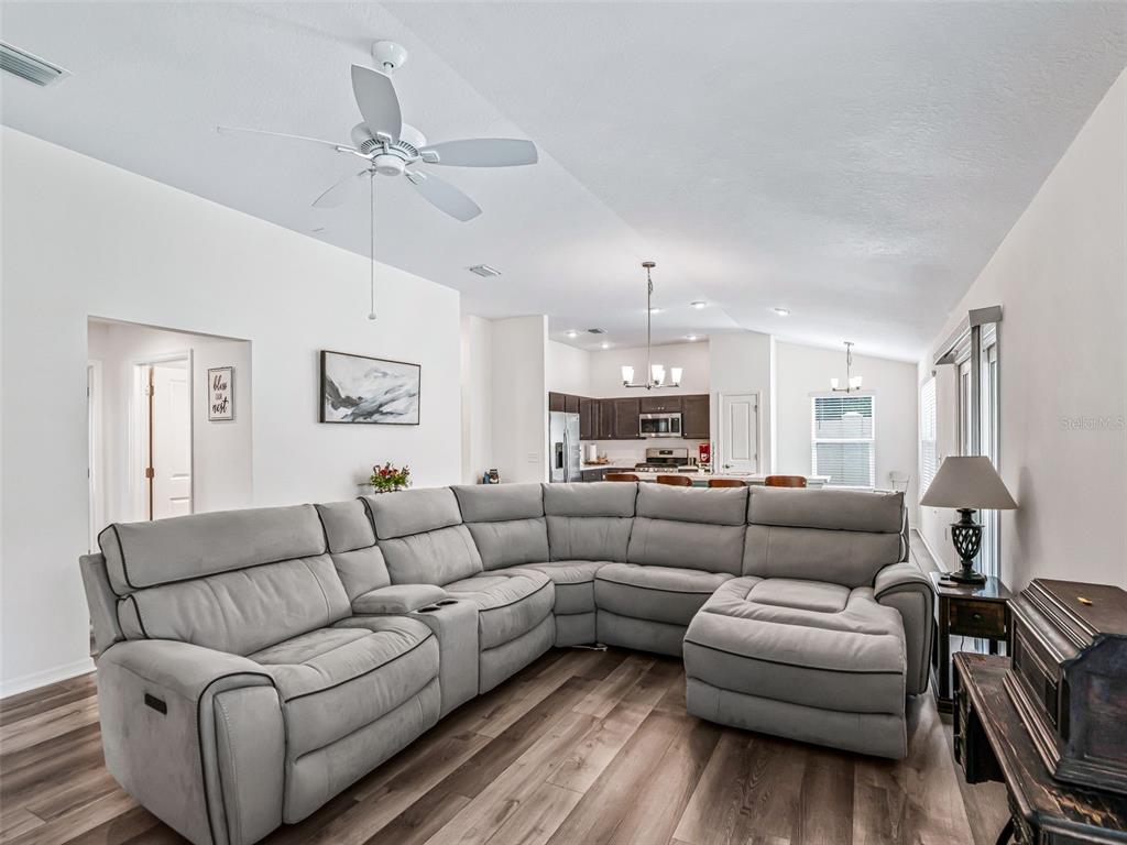 A living room with a large grey sectional sofa, wood-look flooring, and an open kitchen — 4438 CAMEO CIRCLE, THE VILLAGES, FL