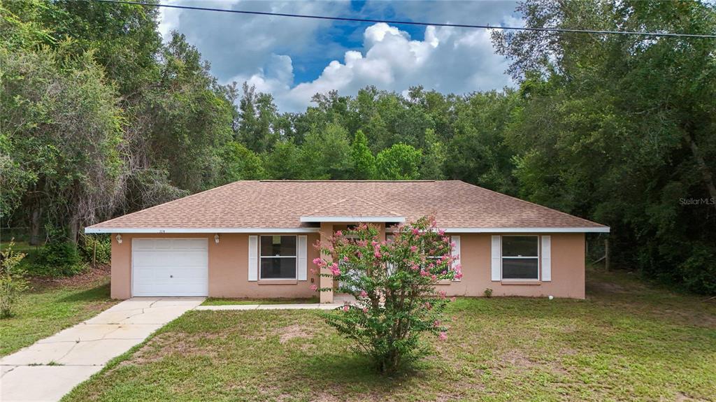 A single-story tan house with a white garage door and a front yard tree — 2210 BLUEGRASS ST, INVERNESS, FL
