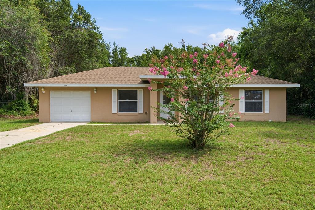 A single-story tan house with a white garage door and a blooming crape myrtle tree — 2210 BLUEGRASS ST, INVERNESS, FL