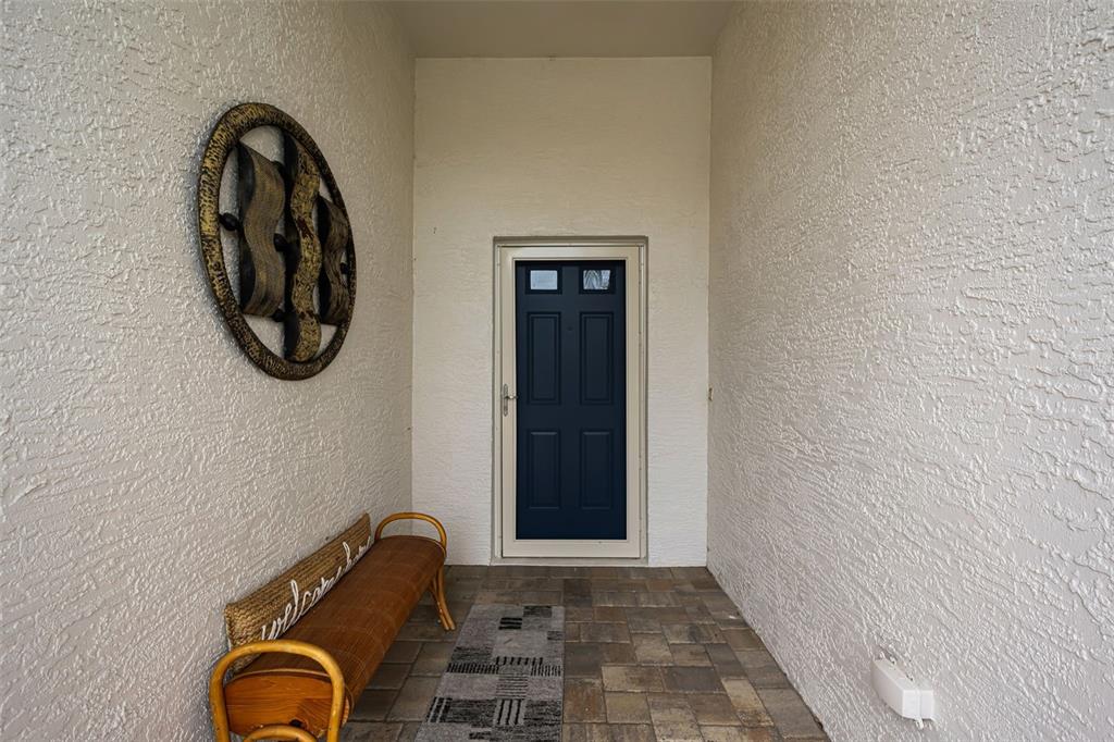 A covered entryway features a dark blue door, a wicker bench, and textured white walls — 1553 ARLINGTON OAKS COURT, NEW PORT RICHEY, FL