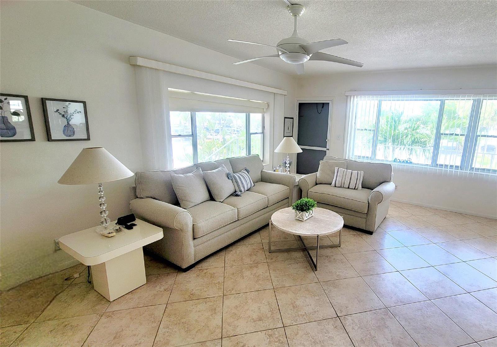 A living room features light-colored furniture, tile flooring, a ceiling fan, and large windows — 3200 NE 7th Court # 208a, Pompano Beach FL 33062, POMPANO BEACH, FL