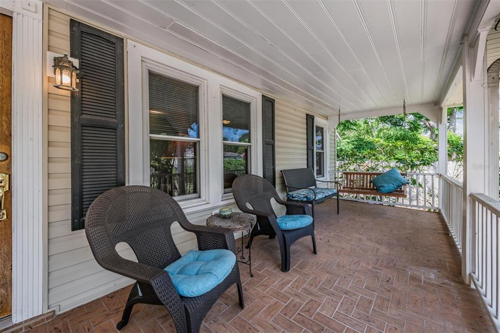 A covered porch features a herringbone brick floor, outdoor seating, and a hanging porch swing — 1618 OTTAWA ROAD, CLEARWATER, FL