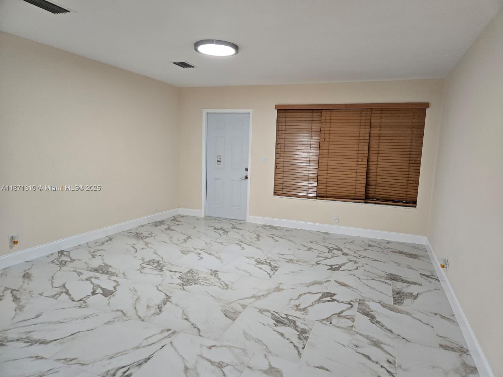 An empty room features white marble-patterned tile flooring, beige walls, a white door, and blinds — 6751 Meade St, Hollywood FL 33024, HOLLYWOOD, FL