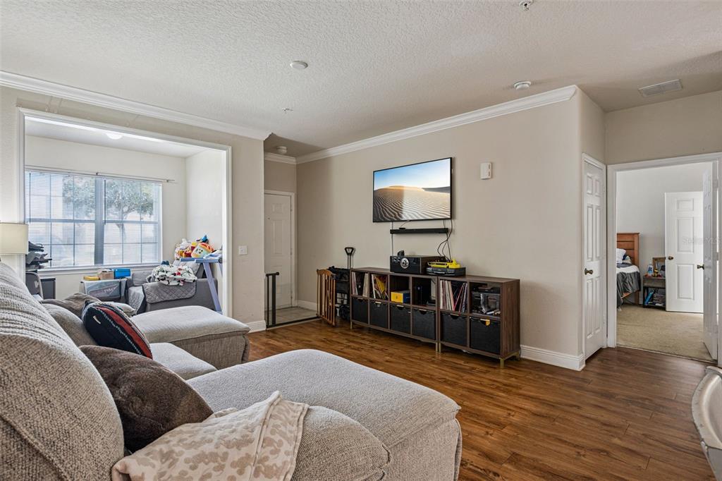 A living room features wood-look flooring, a wall-mounted television, and an adjacent bedroom doorway — 200 SOUTHERN PECAN CIRCLE UNIT 104, WINTER GARDEN, FL