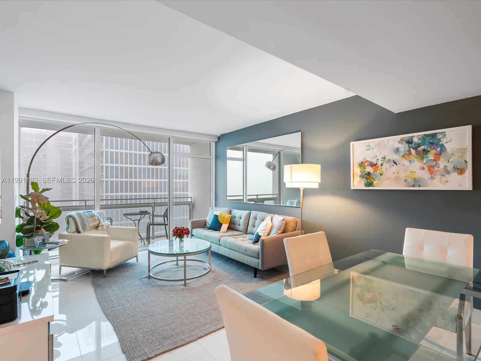 A modern living and dining area features a gray sofa, glass table, and balcony view — 6801 Collins Ave # 1208, Miami Beach FL 33141, MIAMI BEACH, FL